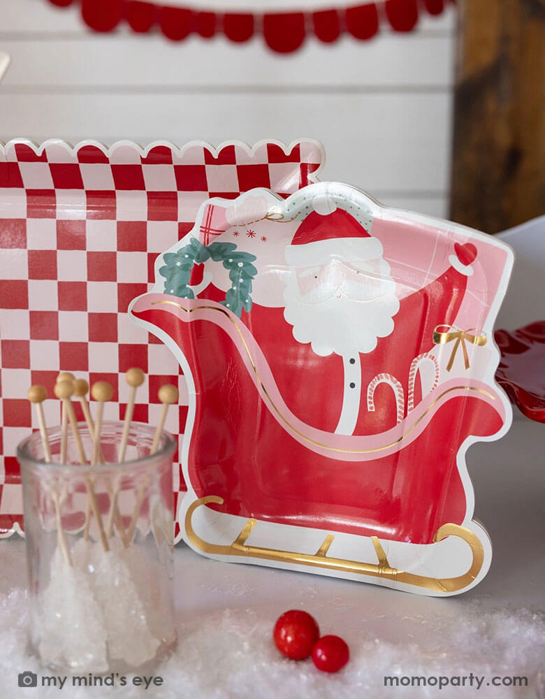 A table shot of Momo Party's 10" Pink and Red Checked Paper Plates next to the Santa on a sleigh shaped plate by My Mind's Eye, with some Holiday candies, gum balls and some artificial snow on the table as decoration. These playful Santa inspired plates add a touch of fun to any table setting. Get ready to serve up some serious style with these quirky and eye-catching plates this Holiday season!