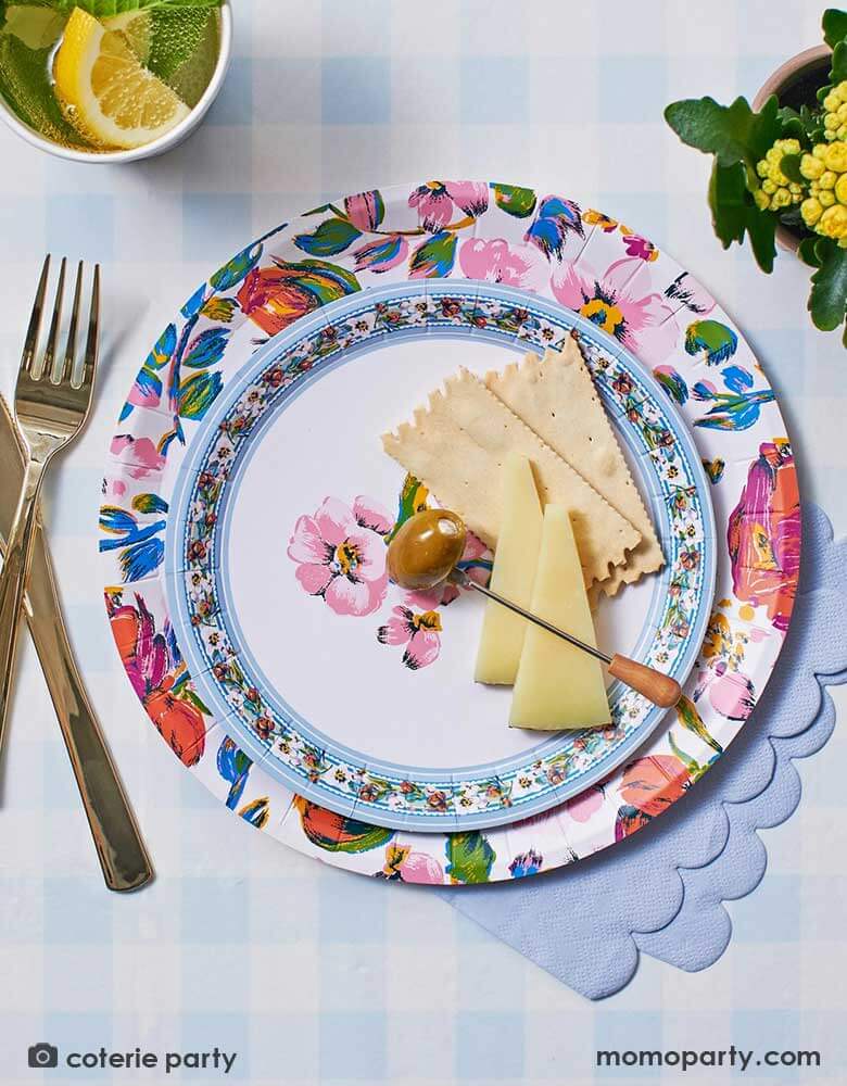 A spring/summer table featuring Momo Party's light blue paper tablecloth by Coterie, along with some floral paper plates, with crackers and cheeses on there, light blue scalloped napkins and some summer flower arrangement and cocktail in the cup, making this a refreshing and stylish tablescape idea this summer.