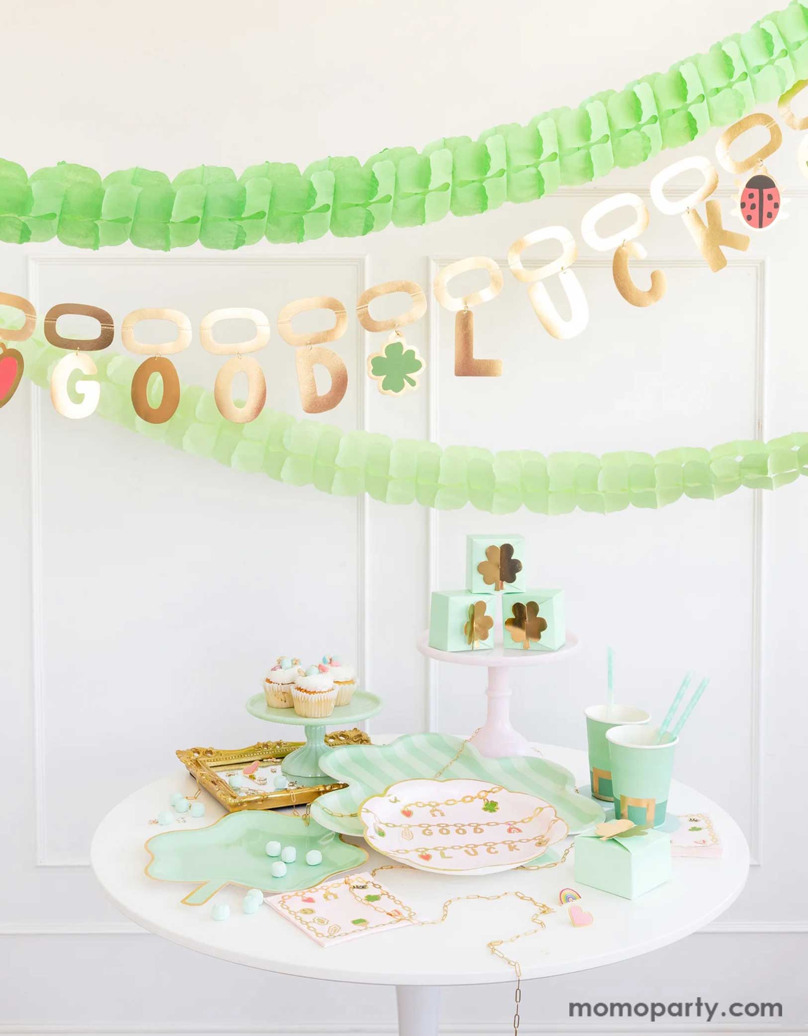 A modern and clean St. Patrick's Day celebration decoration featuring Momo Party's St. Patrick's Day party supplies including shamrock shaped plates, bamboo serving tray, leprechaun shaped party cups, lucky charm scalloped plates and shamrock treat boxes. Above the table hung the good luck charm party banner in gold foil. Along with green tissue honeycomb banner, this creates a charming scene for an adorable St. Patrick's Day celebration for guests of all ages!