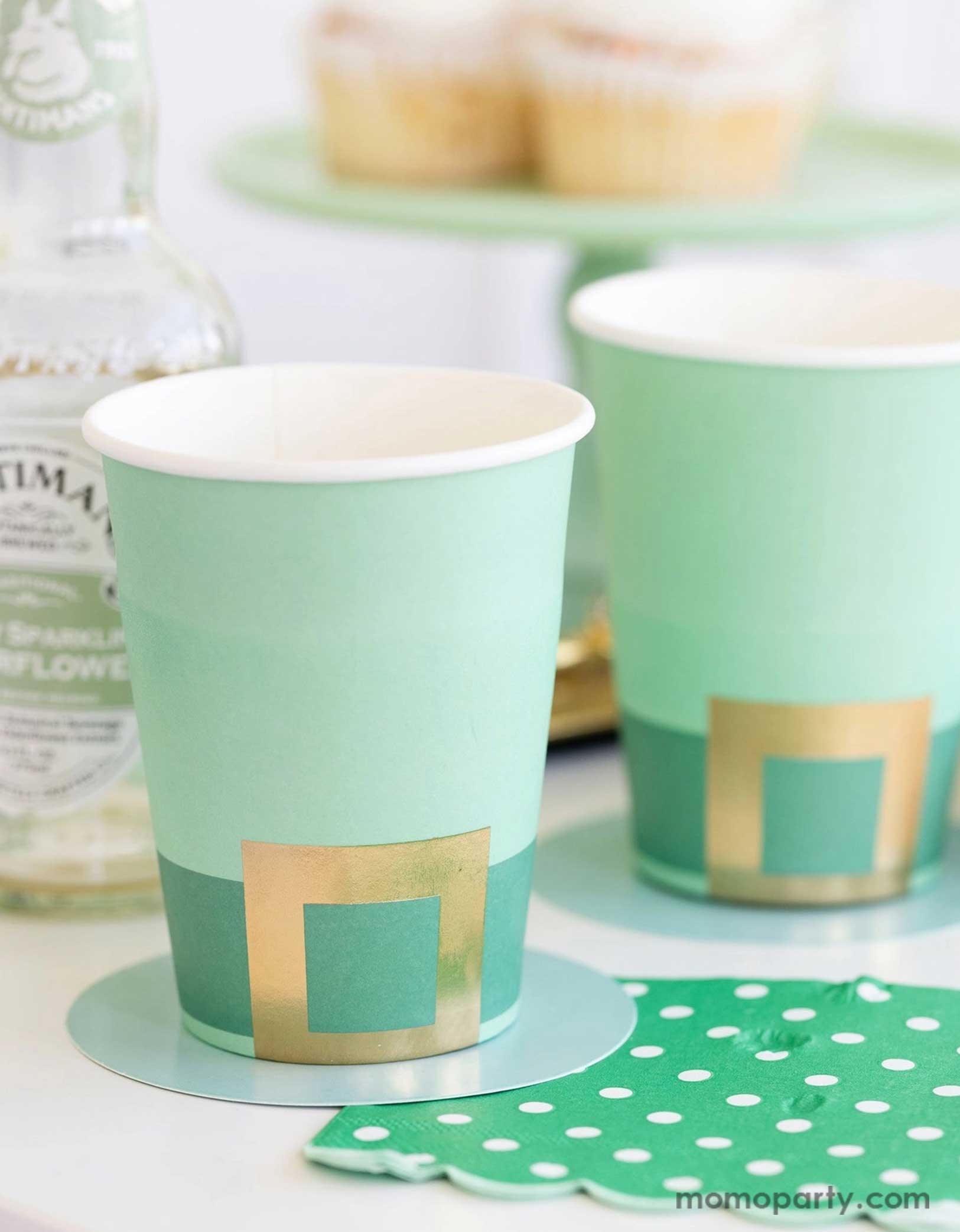 A cheerful St. Patrick's Day party table featuring Momo Party's St. Patrick's Day themed party supplies including the Leprechaun hat party cups and green polka dot napkins. In the back there's a green cake stand with cupcakes on a gold serving tray. With drink bottles next to them making this a fun and festive St. Patrick's Day celebration.