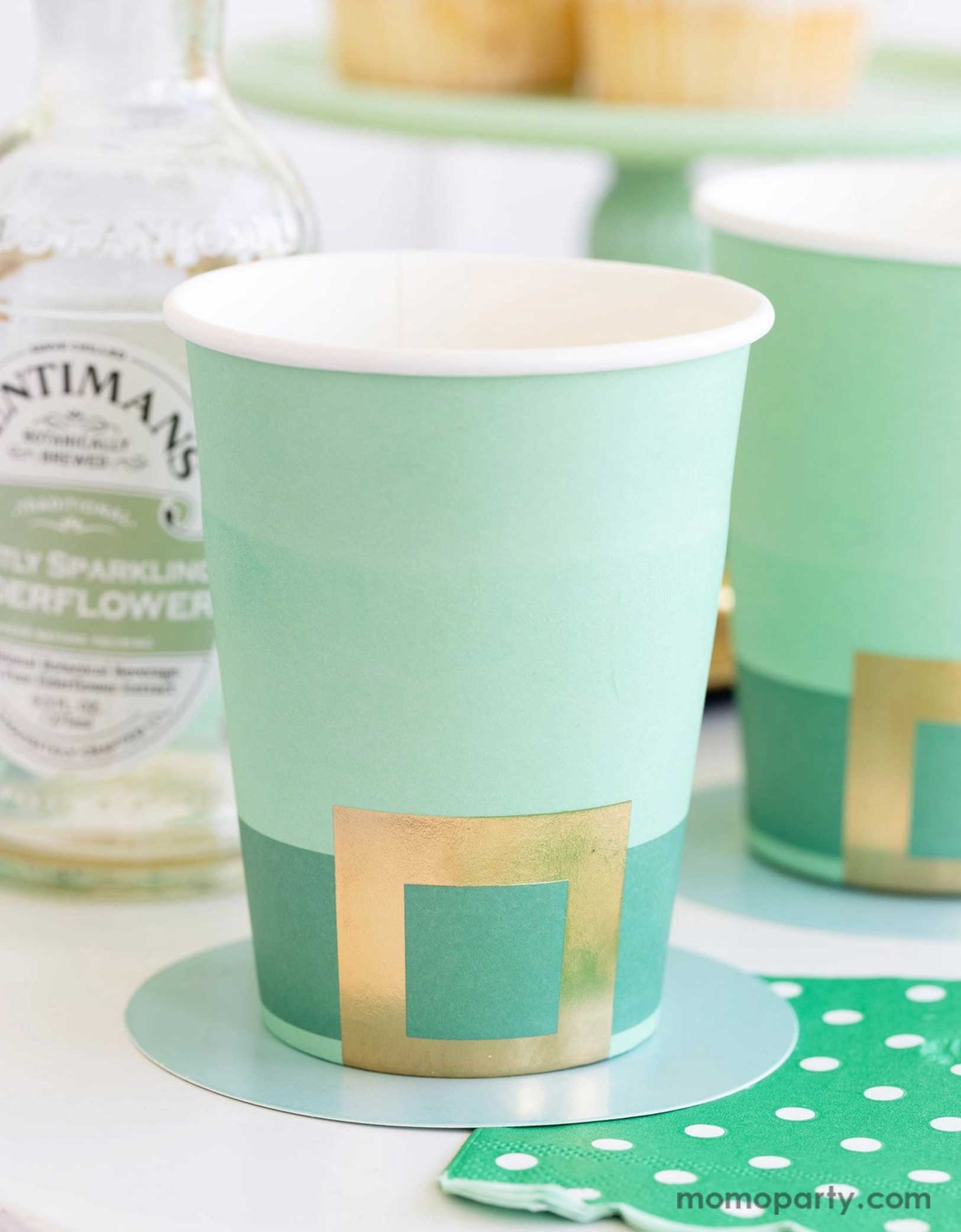 A cheerful St. Patrick's Day party table featuring Momo Party's St. Patrick's Day themed party supplies including the Leprechaun hat party cups and green polka dot napkins. In the back there's a green cake stand with cupcakes on a gold serving tray. With drink bottles next to them making this a fun and festive St. Patrick's Day celebration.