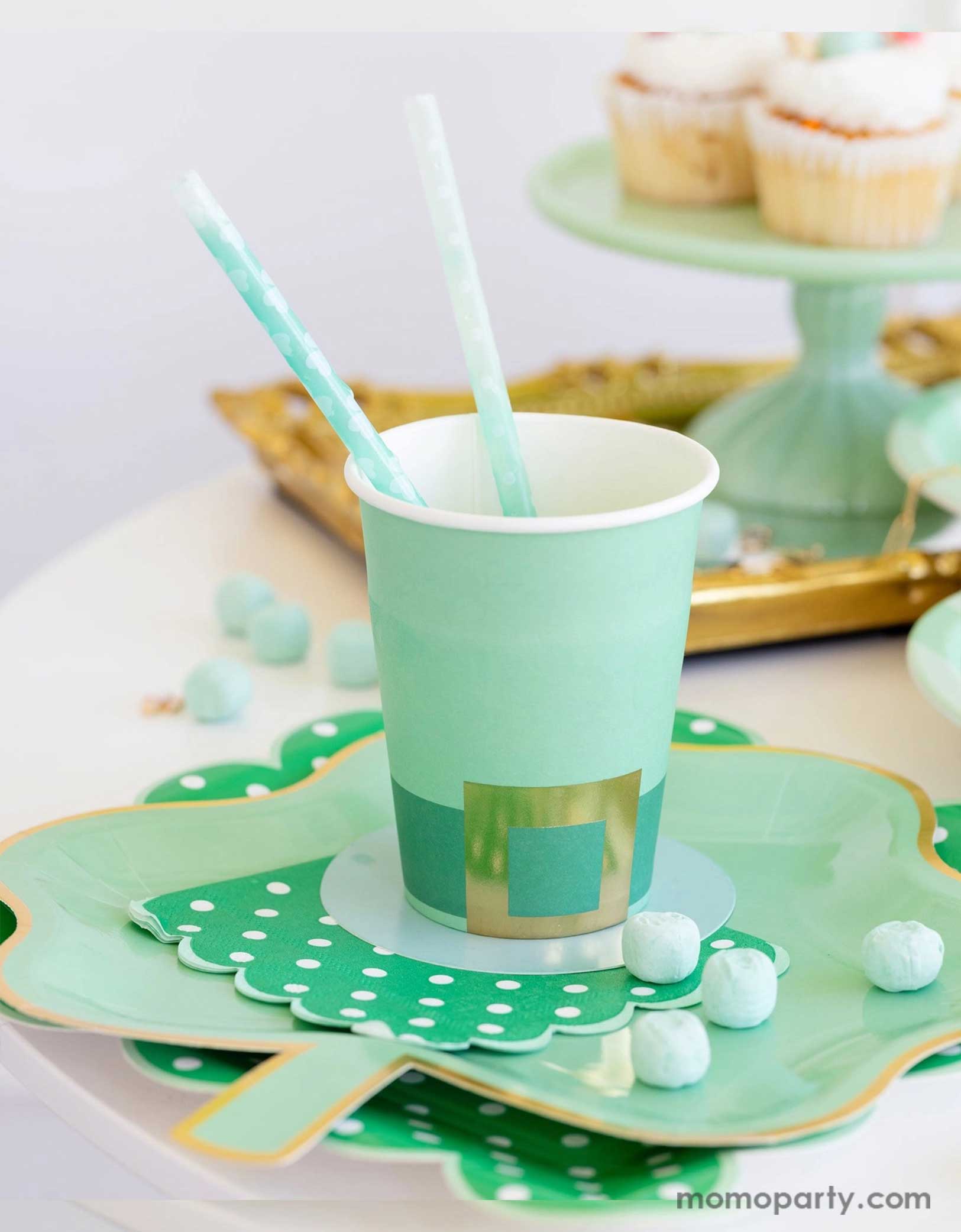 A cheerful St. Patrick's Day party table featuring Momo Party's St. Patrick's Day themed party supplies including the Leprechaun hat party cup, green polka dot napkins, shamrock shaped paper plates, and shamrock reusable straws. In the back there's a green cake stand with cupcakes on a gold serving tray. With green candies around this table makes a great inspo for a fun and festive St. Patrick's Day celebration.