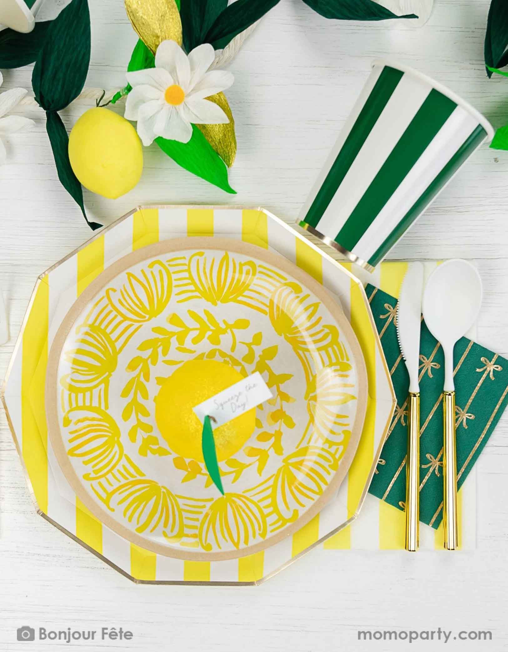 An elegant summer Capri lemon themed party tablescape featuring Momo Party's cabana striped party tableware in yellow and green including table plates, cups and napkins. On the table there's a summer party garland with flowers, greenery, lemons as decorations. 