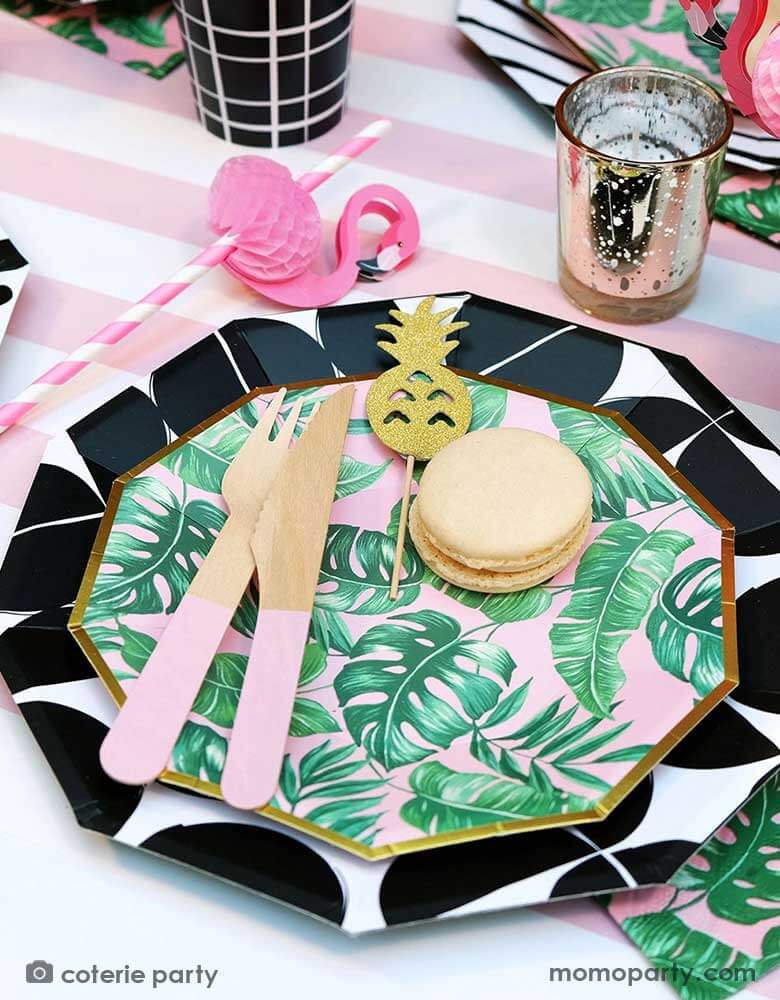 A closeup shot of a modern summer tropical themed party placeset features Momo Party's tropical party themed party supplies including palm leaf plates, napkins, pink wooden utensil set and flamingo honeycomb paper straws in the party cups. All these are on a pink striped table runner, making this a festive yet modern look for a summer party!