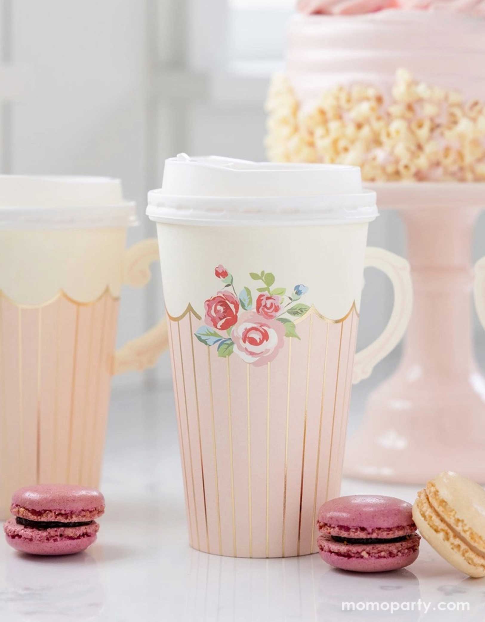 A tea party birthday celebration table set up featuring Momo Party's 12 oz Tea Party Handled Paper Cups by My Mind's Eye. Comes in a set of 8 paper cups, perfect for your tea party on-the-go, these cups feature a clever adhesive handle for easy sipping, with classic rose illustration on the side. Around the paper cups there are some macarons around and a beautiful pale pink cake in the back on a pink cake stand, a perfect setting for a girl's tea party themed birthday celebration. 