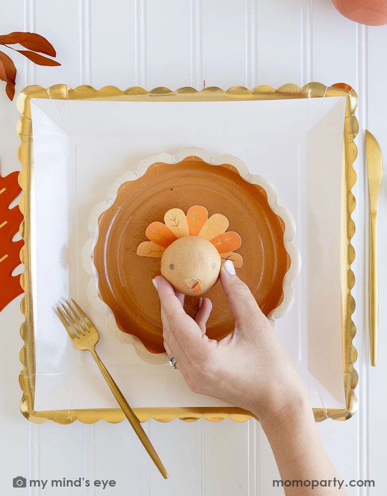 A beautiful and playful Thanksgiving tablescape features Momo Party's Thanksgiving party supplies including gold ruffle large plate and a pie shaped round plate, an oak leaf shaped napkin and a turkey surprise ball. A perfect inspo for a modern Thanksgiving dinner tableset in this harvest season.