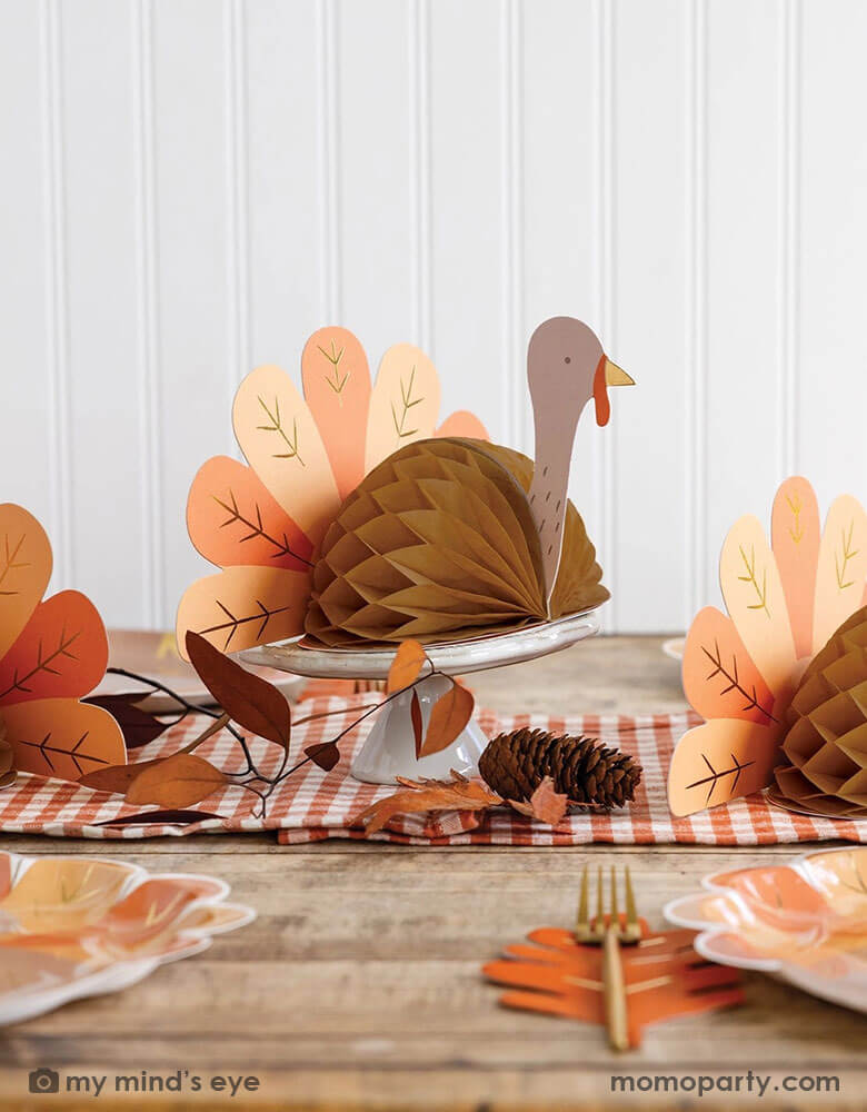 A fall inspired party table features Thanksgiving party supplies by Momo Party including the turkey shaped plates, oak leaf shaped napkins and turkey honeycomb decorations as the centerpiece. With some fall leaf and acorn decorations spread across, this makes a perfect inspo for an autumn gathering or Thanksgiving dinner decor this harvest season.