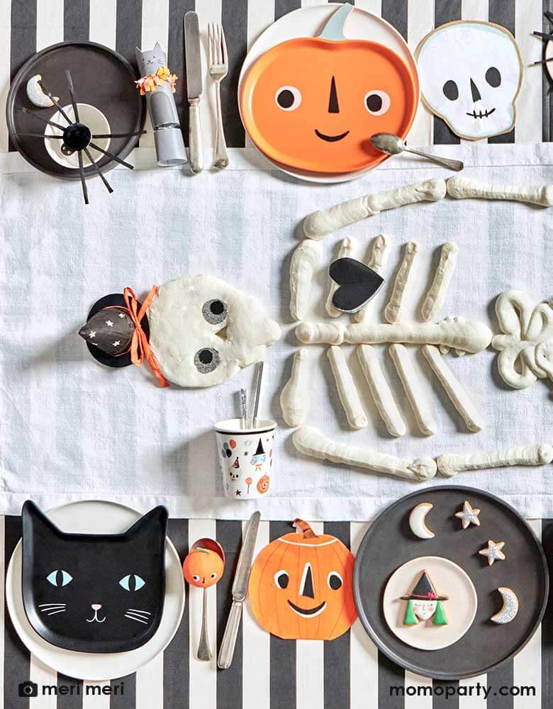 An overhead shot of a Halloween party table featuring Momo Party's modern Halloween tableware including black striped tablecloth, This is Halloween! tableware, napkins, and plates in the classic Halloween icons including pumpkins, skulls, black cats and spiders. In the middle there's skeleton shaped cookies in the middle as the centerpiece making this a modern yet playful tableset for a kid's friendly Halloween table setting ideas.