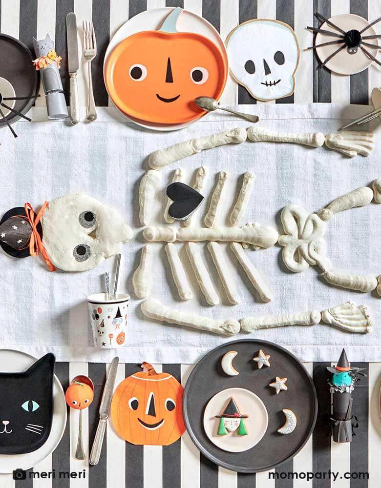 An overhead shot of a Halloween party table featuring Momo Party's modern Halloween tableware including black striped tablecloth, This is Halloween! tableware, napkins, and plates in the classic Halloween icons including pumpkins, skulls, black cats and spiders. In the middle there's skeleton shaped cookies in the middle as the centerpiece making this a modern yet playful tableset for a kid's friendly Halloween table setting ideas.