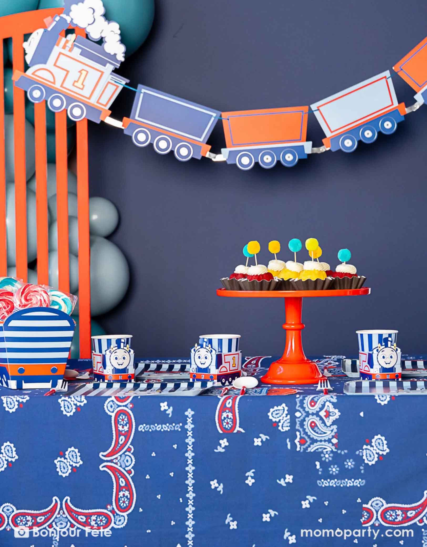 An adorable and festive Thomas the Train and Friends themed train party set up featuring Momo Party's Thomas the Train themed party supplies including party tableware of plates, cups, napkins treat boxes and party garland. The party table is covered with bandana inspired tablecloth to match the theme. In the middle of the table there's a cake stand holding cupcakes in yellow and red colors topped with pom pom toppers. In the back of is a blue balloon garland, making this a cute inspo for kid's train party.