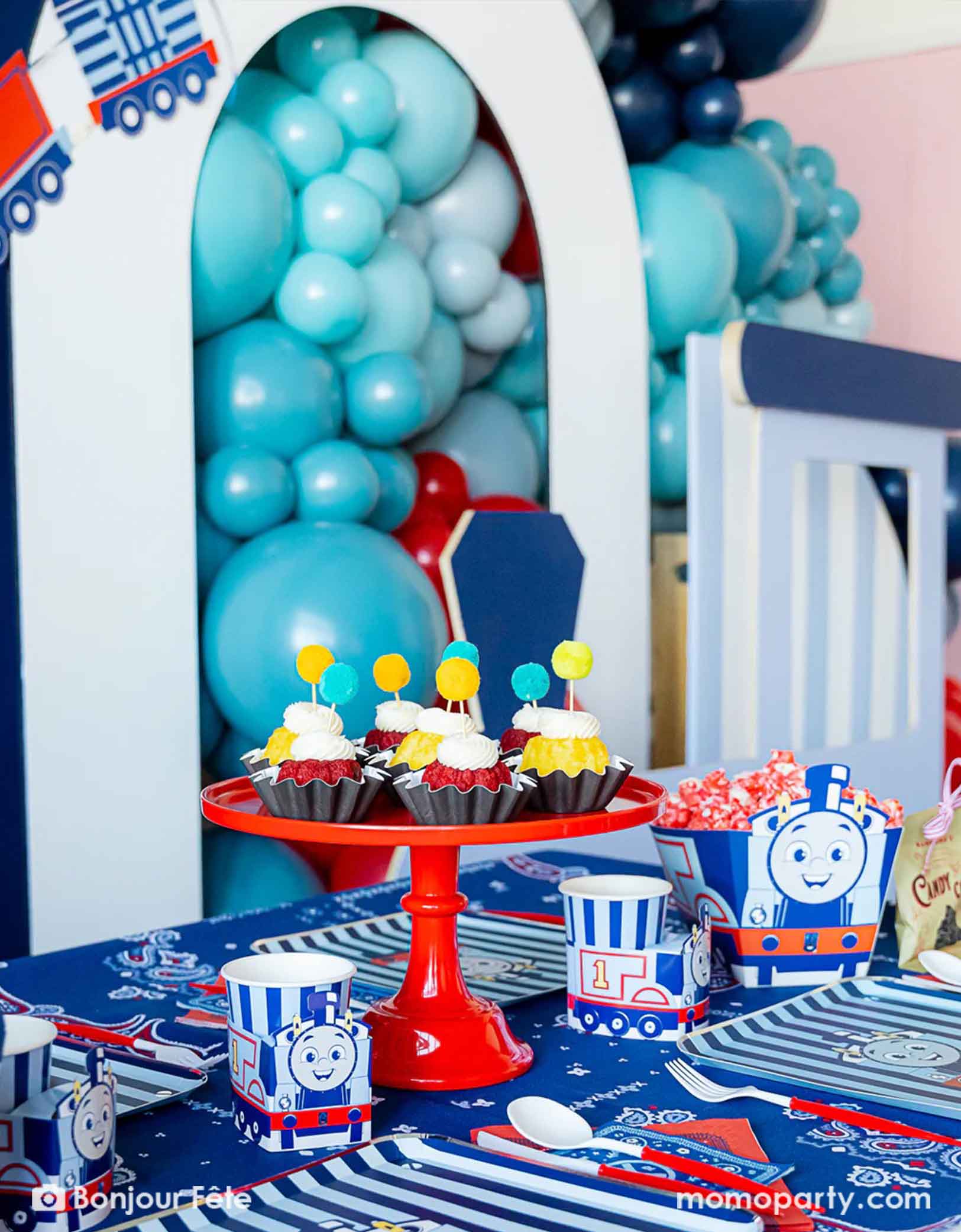 An adorable and festive Thomas the Train and Friends themed train party table featuring Momo Party's Thomas the Train themed party supplies including party tableware of plates, 3D cups, napkins treat boxes. The party table is covered with bandana inspired tablecloth to match the theme. In the middle of the table there's a red cake stand holding cupcakes topped with pom pom toppers. In the back is a blue & red balloon garland with a party garland, making this a perfect inspo for toddler's train party.