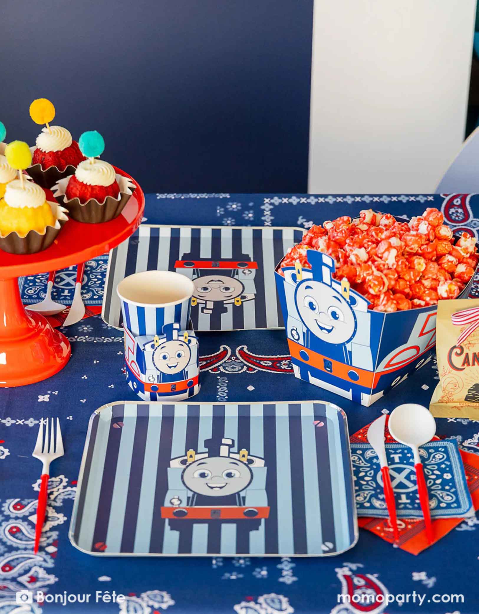 An adorable and festive Thomas the Train and Friends themed train party table featuring Momo Party's Thomas the Train themed party supplies including party tableware of striped plates, 3D cups, napkins and treat boxes. The party table is covered with bandana inspired tablecloth to match the theme. In the middle of the table there's a red cake stand holding cupcakes topped with pom pom toppers. In the back is a blue & red balloon garland, making this a perfect inspo for toddler's train themed birthday party.