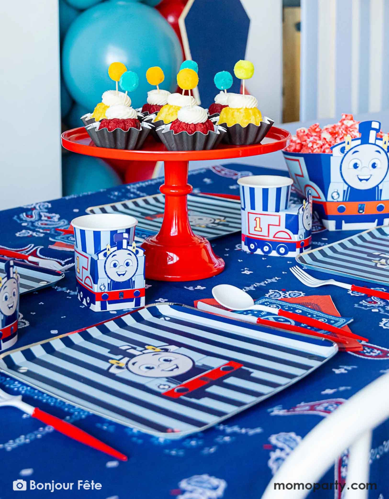 An adorable and festive Thomas the Train and Friends themed train party table featuring Momo Party's Thomas the Train themed party supplies including party tableware of plates, 3D cups, napkins treat boxes. The party table is covered with bandana inspired tablecloth to match the theme. In the middle of the table there's a red cake stand holding cupcakes topped with pom pom toppers. In the back is a blue & red balloon garland, making this a perfect inspo for toddler's train themed birthday party.