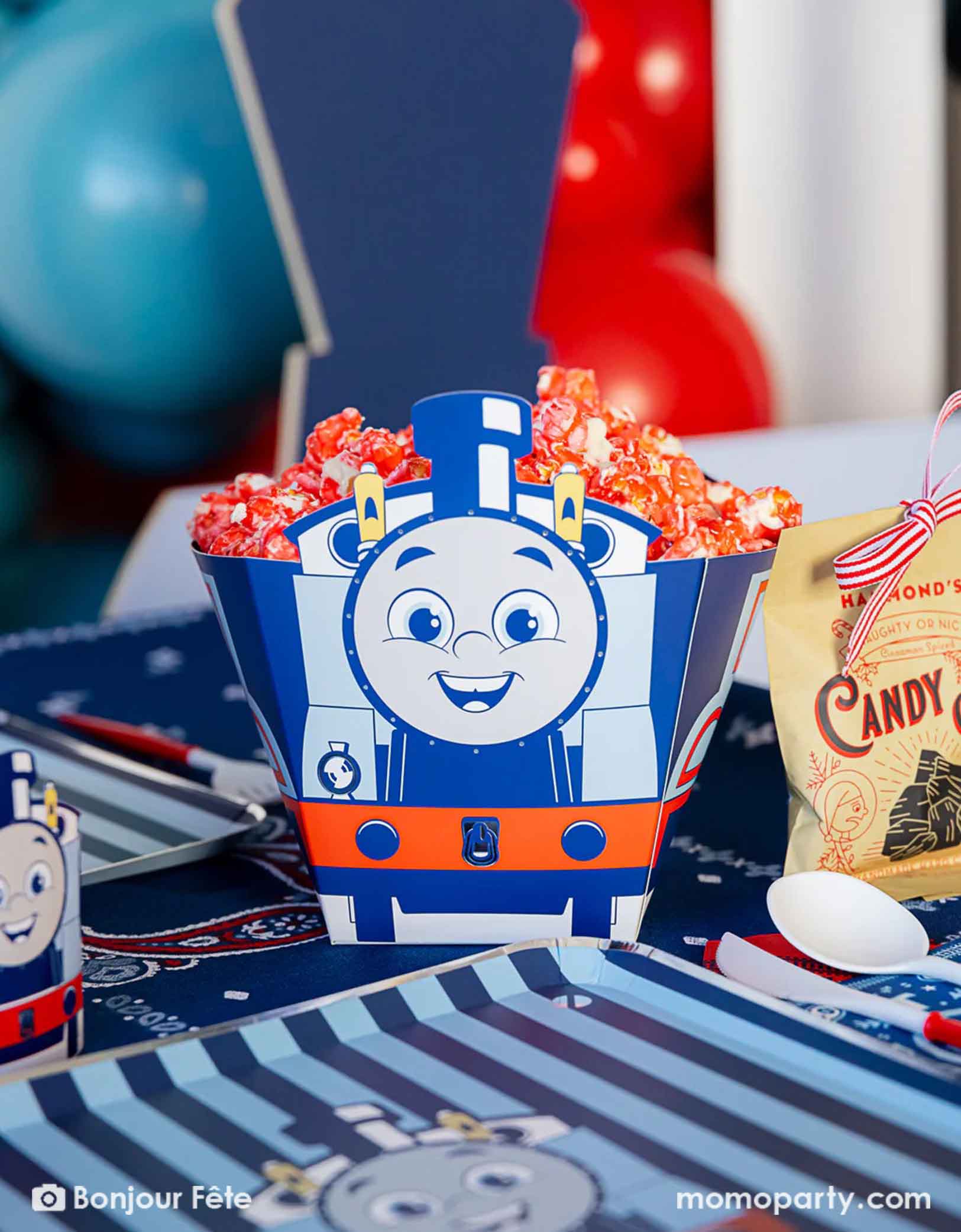 A close-up of the shot of Thomas the Train and Friends shaped treat box filled with red popcorn in a festive Thomas the Train and Friends themed train party. On the table there are various Thomas the Train themed party supplies from Momo Party including party tableware of plates, 3D cups, napkins treat boxes. The party table is covered with bandana inspired tablecloth to match the theme. In the back is a blue & red balloon cloud, making this a perfect inspo for a toddler's train birthday celebration.