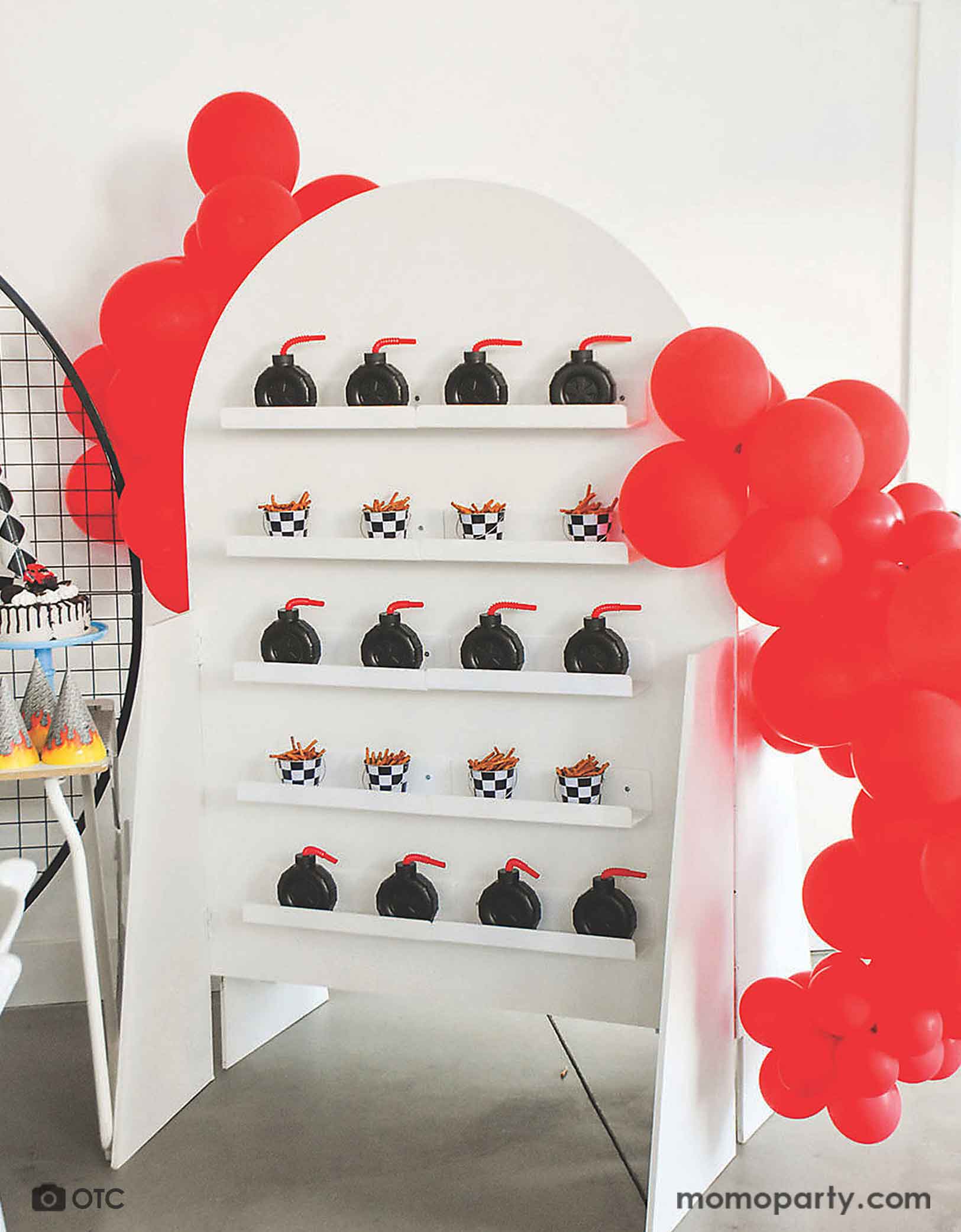 A kid's monster truck themed birthday party favor wall filled with Momo Party's 12 oz 4" tire truck shaped cups and mini checkered board buckets with pretzels. Around the wall, there are two red balloon garlands, making this a modern and clean set up for a fun kid's monster truck car themed birthday celebration!