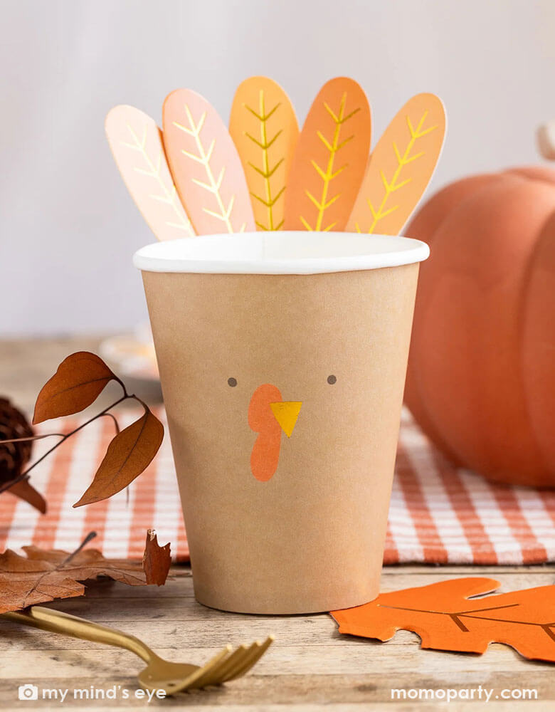 A Thanksgiving party table shot featuring Momo Party's 8 oz Harvest Turkey Paper Party Cup by My Mind's Eye. On the table there are some fall leaves decoration, an oak leaf shaped napkin, a pumpkin and an orange gingham checkered table runner, this cozy table setting makes a great inspiration for this harvest season. 