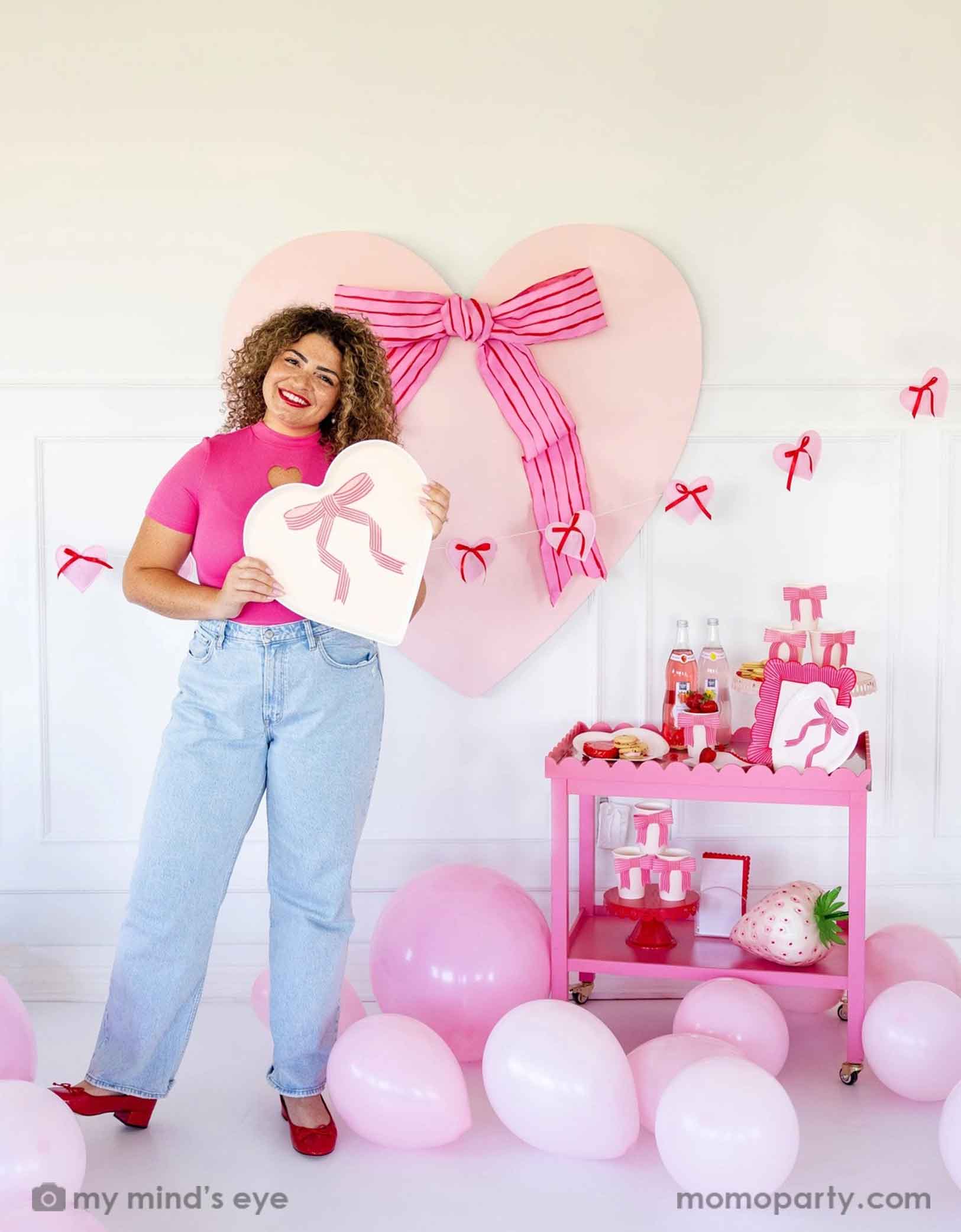 A lady with a hot pink top with red lipstick and red high heels holding Momo Party's 15" x 15"  Bow Heart Tray by My Mind's Eye. She is standing next to a pink cart filled with Valentine's Day party supplies including plates, cups, napkins, along with drinks and snacks. In her back there's a giant pink heart shaped decoration with ribbon bow on the wall. With a floor of pink balloons, this makes a great scene for a sweet Valentine's Day celebration.