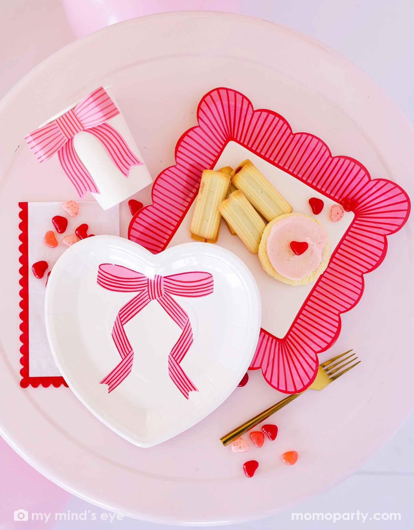 A overhead shot of Momo Party's bow heart Valentine's Day party collection including light pink heart shaped plate, bow party cup, striped scallop edged plate and scallop napkins. Around the tableware there are some heart shaped candies, cookies and some Valentine's Day themed snacks.