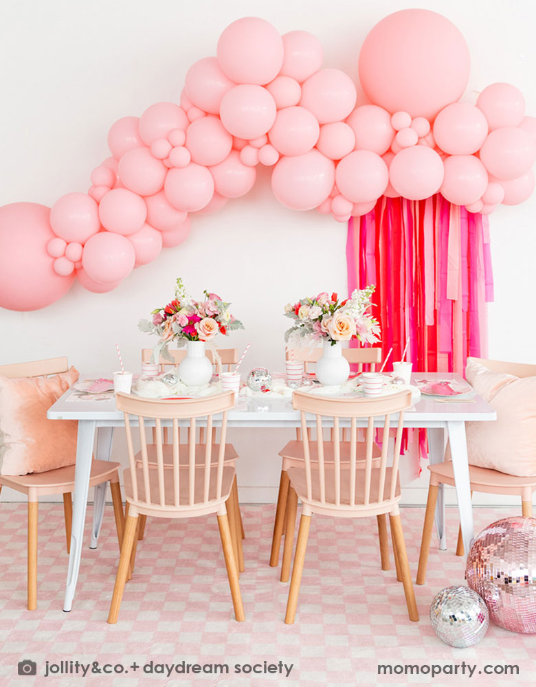 A beautiful Valentine's Day party set up featuring a gorgeous pink balloon garland adorned with rose pink streamer. On the party table there are Momo Party's red striped tableware including cups, plates, napkins, along with some pretty pink and red flower arrangement. Along with light pink wooden chairs with pink pillows, pink checkered rug and large pink disco balls, this makes a great inpo for a darling Valentine's Day celebration for girl's gatherings. 