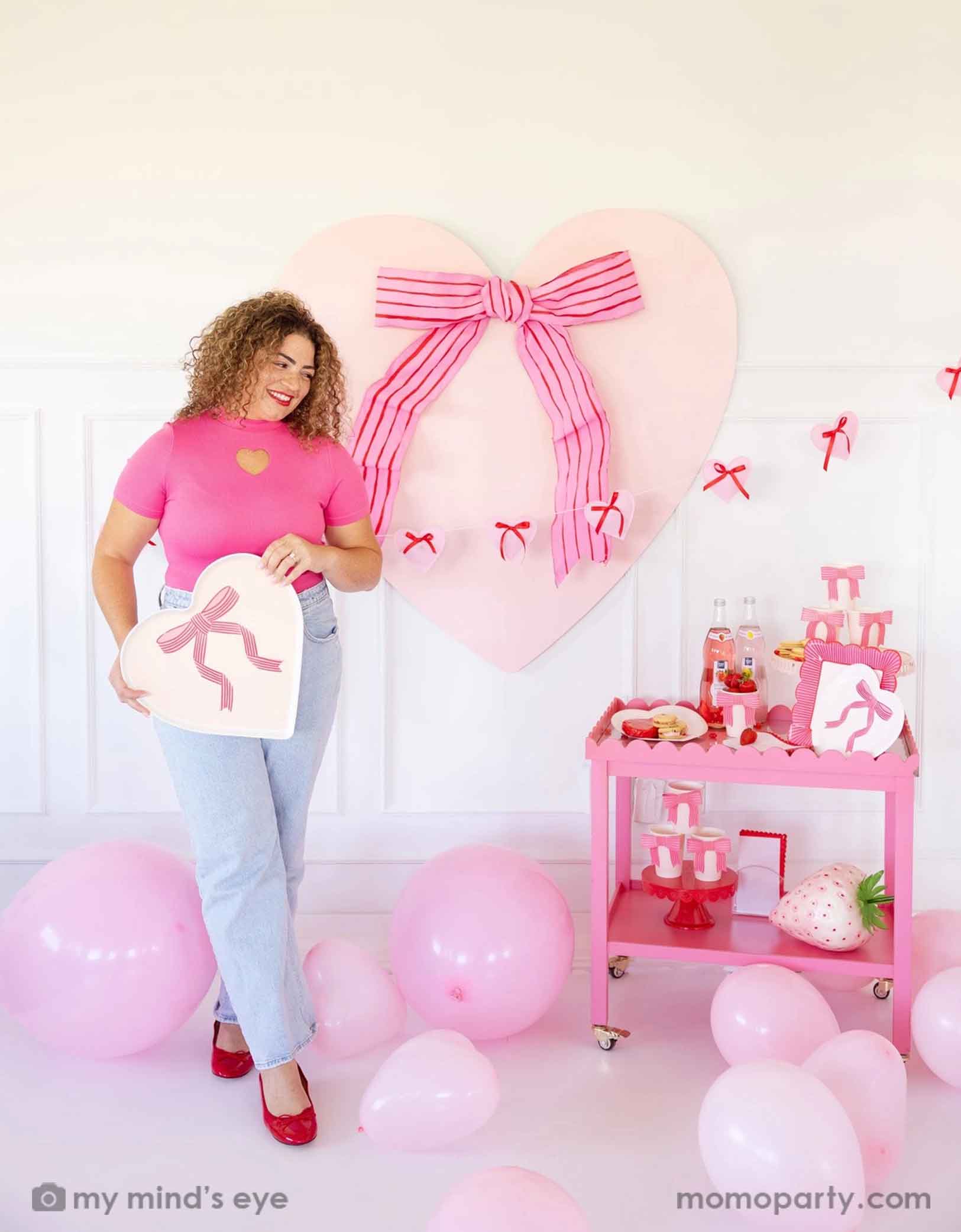 A lady with a hot pink top with red lipstick and red high heels holding Momo Party's 15" x 15"  Bow Heart Tray by My Mind's Eye. She is standing next to a pink cart filled with Valentine's Day party supplies including plates, cups, napkins, along with drinks and snacks. In her back there's a giant pink heart shaped decoration with ribbon bow on the wall. With a floor of pink balloons, this makes a great scene for a sweet Valentine's Day celebration.