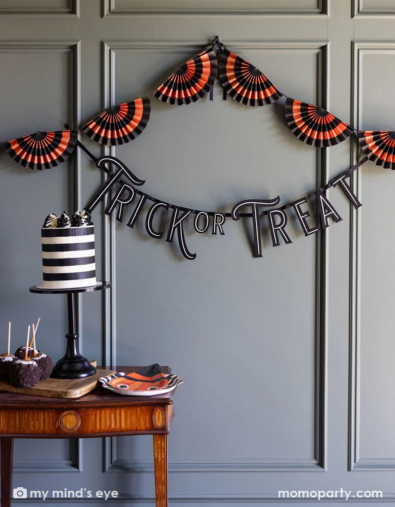 A room decorated with Momo Party's vintage Halloween themed party supplies and decorations by My Mind's Eye including the 4ft Trick or Treat felt banner, black and orange paper bunting banner on the wall. In the front there's a vintage desk with a black striped cake and some caramel apples dipped with Oreo chocolate crumbs. A stack of vintage Halloween pumpkin shaped paper plates are on the side for serving, making this a unique and spooky inspo for this Halloween season.