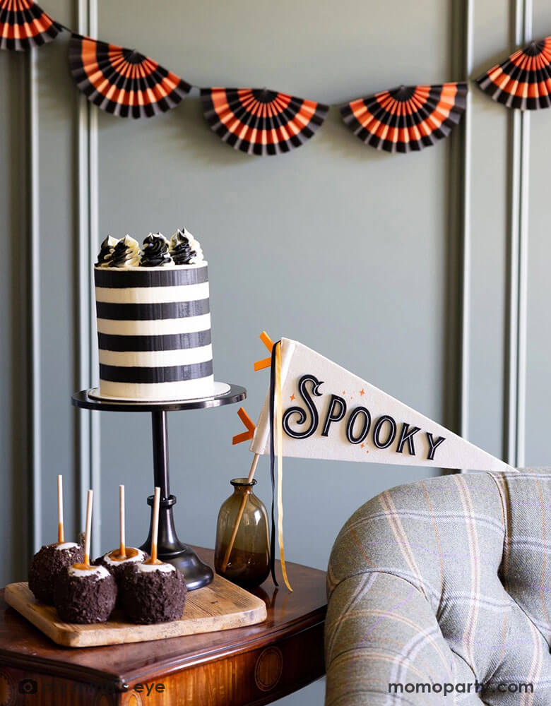 A room decorated with Momo Party's Vintage Halloween themed party supplies and decorations by My Mind's Eye including the the black and orange paper bunting banner on the wall. In the front there's a vintage desk with a black striped cake and some caramel apples dipped with Oreo chocolate crumbs, with the Spooky felt party pennant in a small vase. Making this a unique and spooky inspo for this Halloween season.