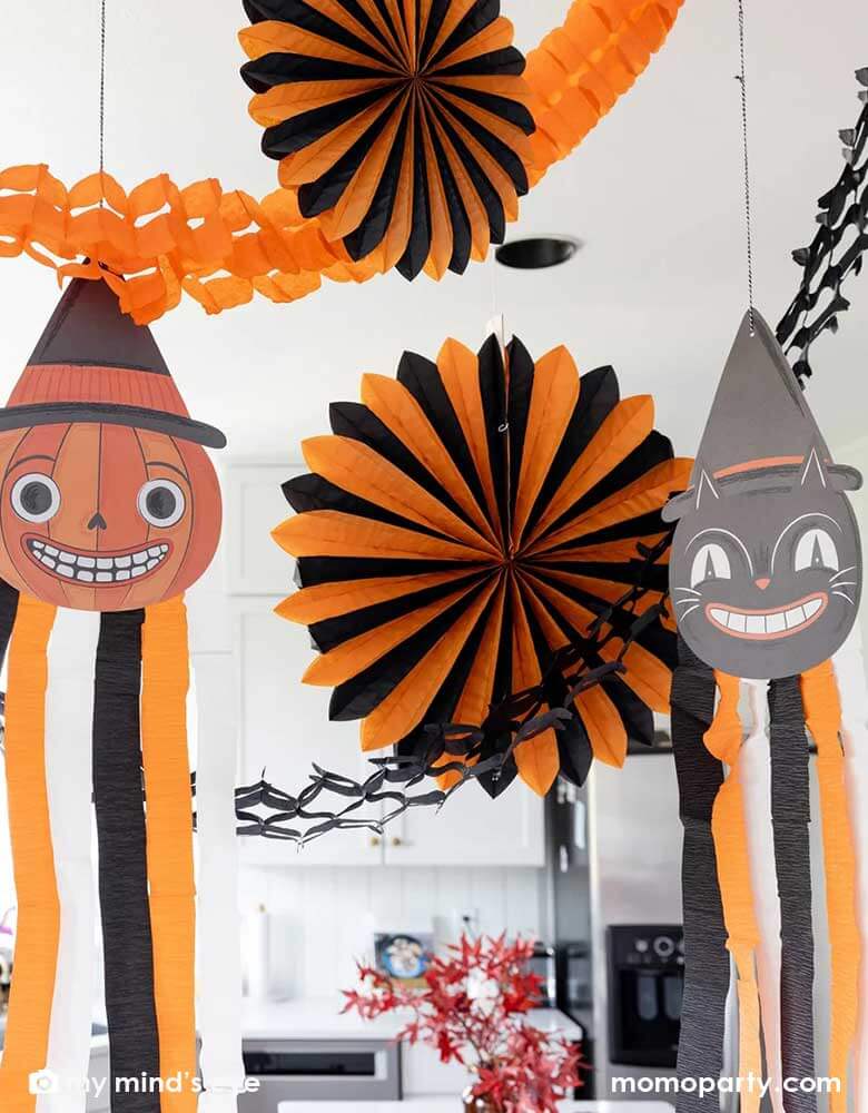 A close-up shot of a modern kitchen that's decorated with vintage inspired Halloween party supplies form Momo Party including the Halloween hanging characters of a black cat and a pumpkin, which around them hung the oversized orange and black paper fans and tissue decorations. Making this a eerie yet fun decoration that sets a festive scene for your Halloween celebration this spooky season.