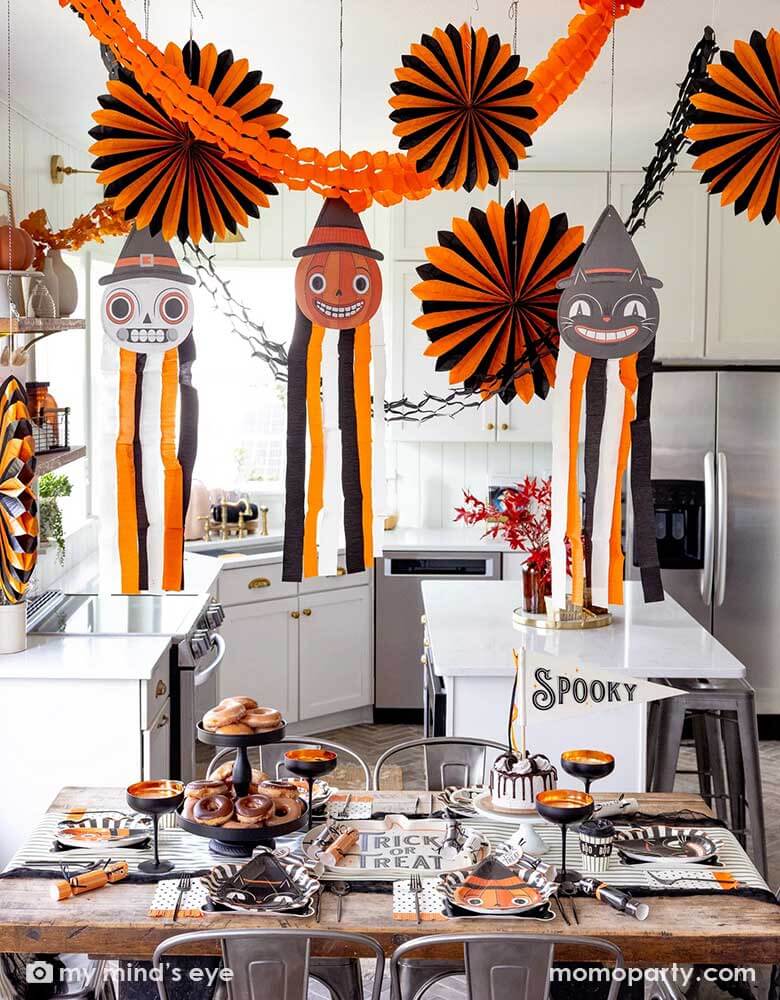 A modern kitchen that's decorated with vintage inspired Halloween party supplies form Momo Party including the Halloween hanging characters of a black cat, a pumpkin and a skull, which around them hung the oversized orange and black paper fans and tissue decorations. On the table there are matching tableware including party plates, napkins, cups and party crackers in the vintage style. With the "Spooky" felt party pennant, cakes and donuts, this creates a festive vibe for a fun Halloween celebration. 