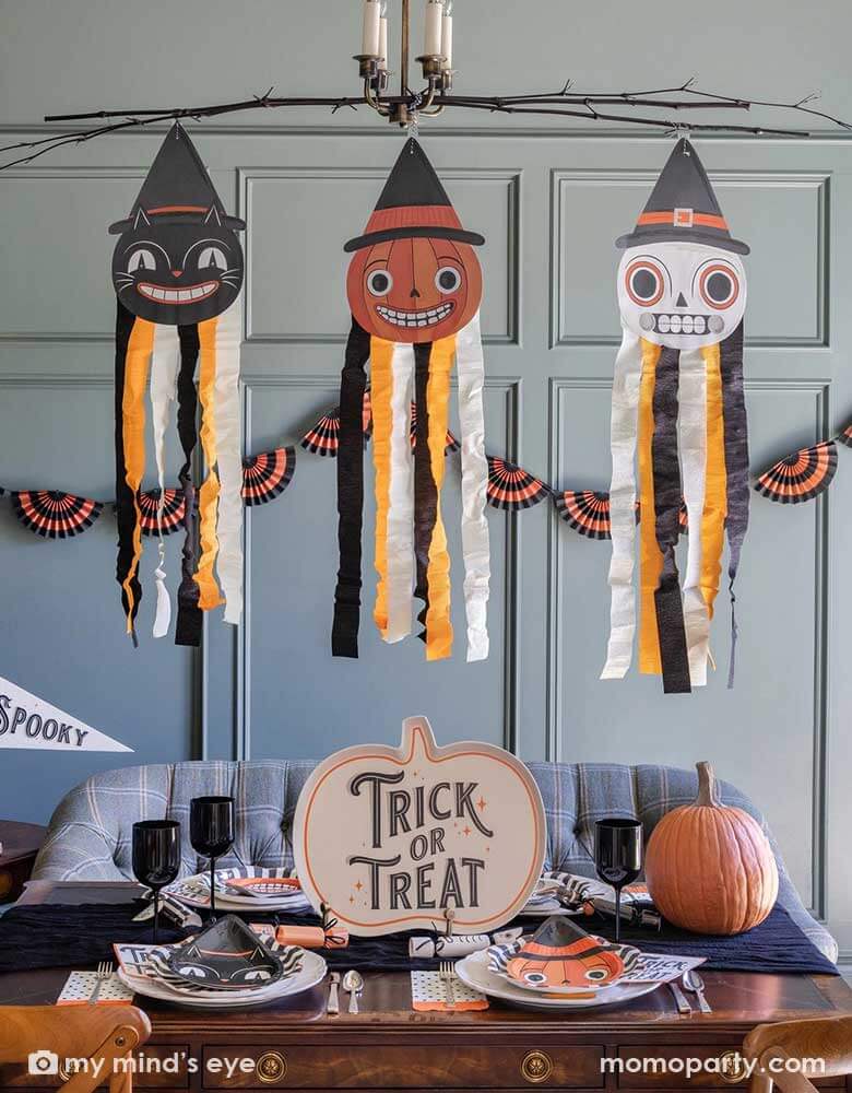 A Halloween party set up featuring vintage Halloween party supplies and decorations from Momo Party including the giant vintage Halloween character hanging decoration streamers and the black and orange bunting banner on the wall as the backdrop. On the table, there's a huge pumpkin shaped bamboo serving plate that says trick or treat on it. With vintage Halloween themed plates, napkins and glasses. This makes a great inspo for a fun Halloween gathering at home.