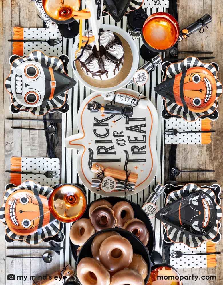 A overhead shot of a festive Halloween party table featuring Momo Party's vintage Halloween character shaped plates on the black starburst dinner plates and night sky scalloped plates, plus black dotted napkins with orange fringe. On the table there are some vintage Halloween party crackers on "Trick or Treat" pumpkin shaped bamboo serving tray on the black and white striped table runner. With delicious treats like cakes, donuts and apple cider, it's ready for a spooky fun Halloween celebration!