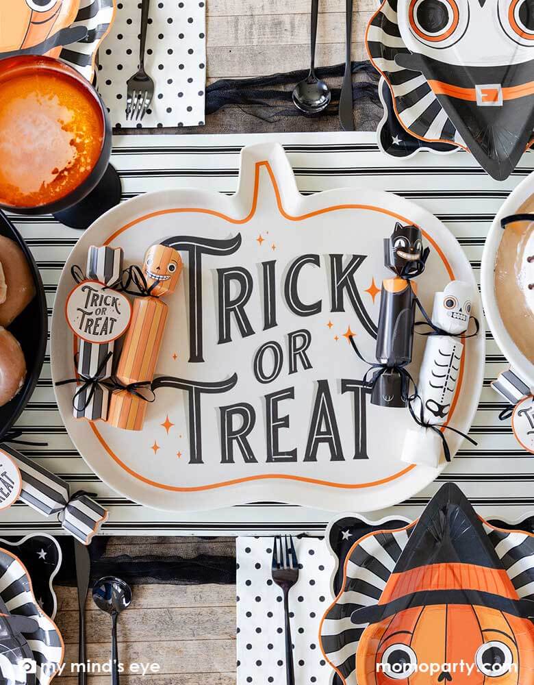 A close up of a festive Halloween party table featuring Momo Party's vintage Halloween party supplies including Halloween party crackers in the designs of a pumpkin, a black at and a skeleton on  the"Trick or Treat" pumpkin shaped bamboo serving tray on the black and white striped table runner. Around the centerpiece were the Halloween character shaped plates layered with black starburst plates. With delicious treats like cakes, donuts and apple cider, it's ready for a spooky fun Halloween celebration!