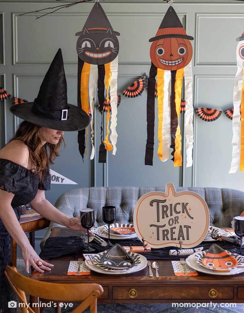 A lady dressed as witch setting up a Halloween party table in a vintage inspired Halloween party featuring Halloween party supplies and decorations from Momo Party including the giant vintage Halloween character hanging decoration streamers and the black and orange bunting banner on the wall as the backdrop. On the table, there's a huge pumpkin shaped bamboo serving plate that says trick or treat on it. With vintage Halloween themed plates, napkins and glasses.