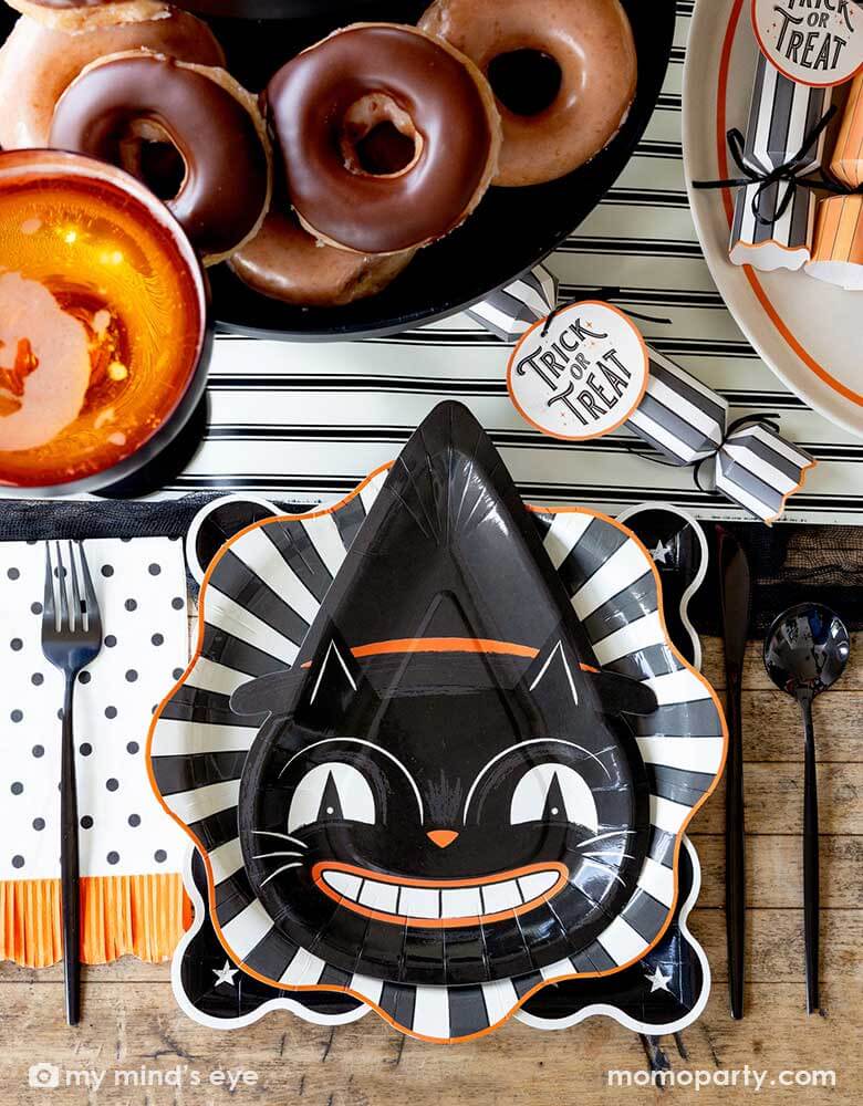 A vintage Halloween party table setting featuring vintage inspired Halloween party supplies from Momo Party including the vintage Halloween black cat shaped plate on the black starburst dinner plate and night sky scalloped plate. On the table there are some vintage Halloween party crackers on a black and white striped table runner. With delicious treats like donuts and apple cider, this festive table is ready for a spooky fun Halloween celebration!