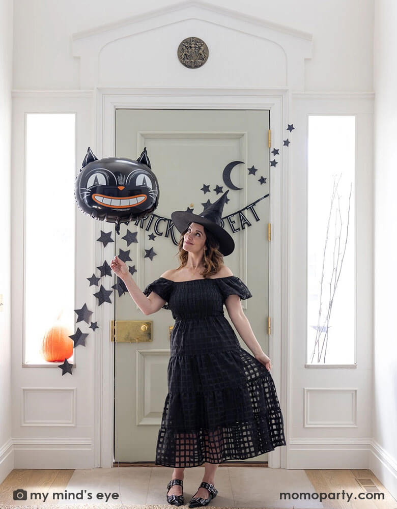 A lady dressed as a Halloween witch holding Momo Party's 24" black cat shaped foil balloon at the doorway decorated with Momo Party's 4ft Trick or Treat felt banner and glittered stars and moon wall paper decorations by My Mind's Eye. Behind the door through the side windows you can see a pumpkin and some spooky branches as the decorations, making this a perfect Halloween home decor idea for this spooky season!