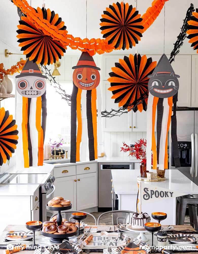 A modern kitchen that's decorated with vintage inspired Halloween party supplies form Momo Party including the Halloween hanging characters of a black cat, a pumpkin and a skull, which around them hung the oversized orange and black paper fans and tissue decorations. On the table there are matching tableware including party plates, napkins, cups and party crackers in the vintage style. With the "Spooky" felt party pennant, cakes and donuts, this creates a festive vibe for a fun Halloween celebration. 