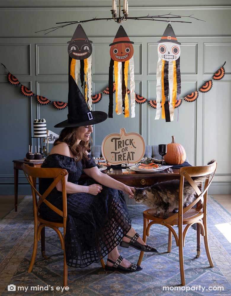 A lady dressed as witch playing with her cat sitting next to her in Halloween party set up featuring vintage Halloween party supplies and decorations from Momo Party including the giant vintage Halloween character hanging decoration streamers and the black and orange bunting banner on the wall as the backdrop. On the table, there's a huge pumpkin shaped bamboo serving plate that says trick or treat on it. With vintage Halloween themed plates, napkins and glasses.