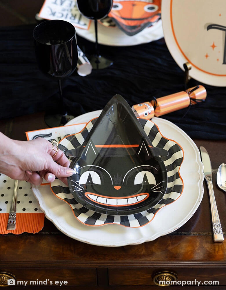 A Halloween party table setting featuring vintage inspired Halloween party supplies from Momo Party. The hostess is placing the vintage Halloween black cat shaped plate onto the black starburst large plates and there is the vintage Halloween trick or treat small napkin next to the plates. On the table there are some vintage Halloween character party crackers in the design of a skeleton, a black cat and a pumpkin. Making this a perfect inspo for a festive and spooky Halloween celebration!