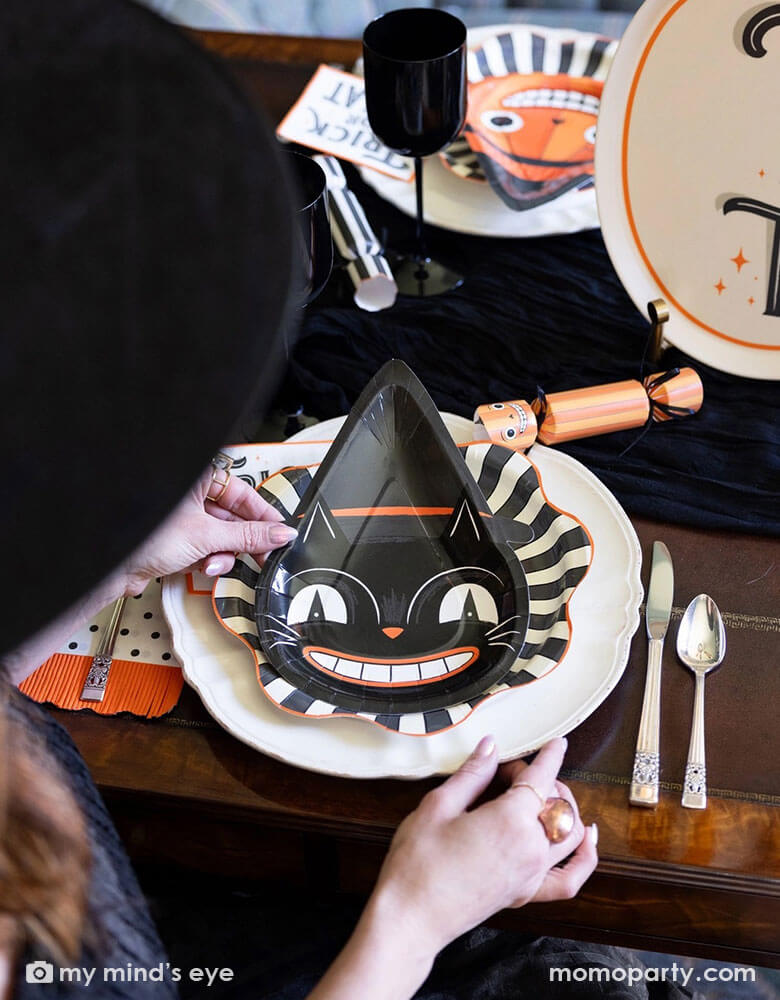 A Halloween party table setting featuring vintage inspired Halloween party supplies from Momo Party. The hostess is placing the vintage Halloween black cat shaped plate onto the black starburst large plates and there is the vintage Halloween trick or treat small napkin next to the plates. On the table there are some vintage Halloween character party crackers in the design of a skeleton, a black cat and a pumpkin. Making this a perfect inspo for a festive and spooky Halloween celebration!