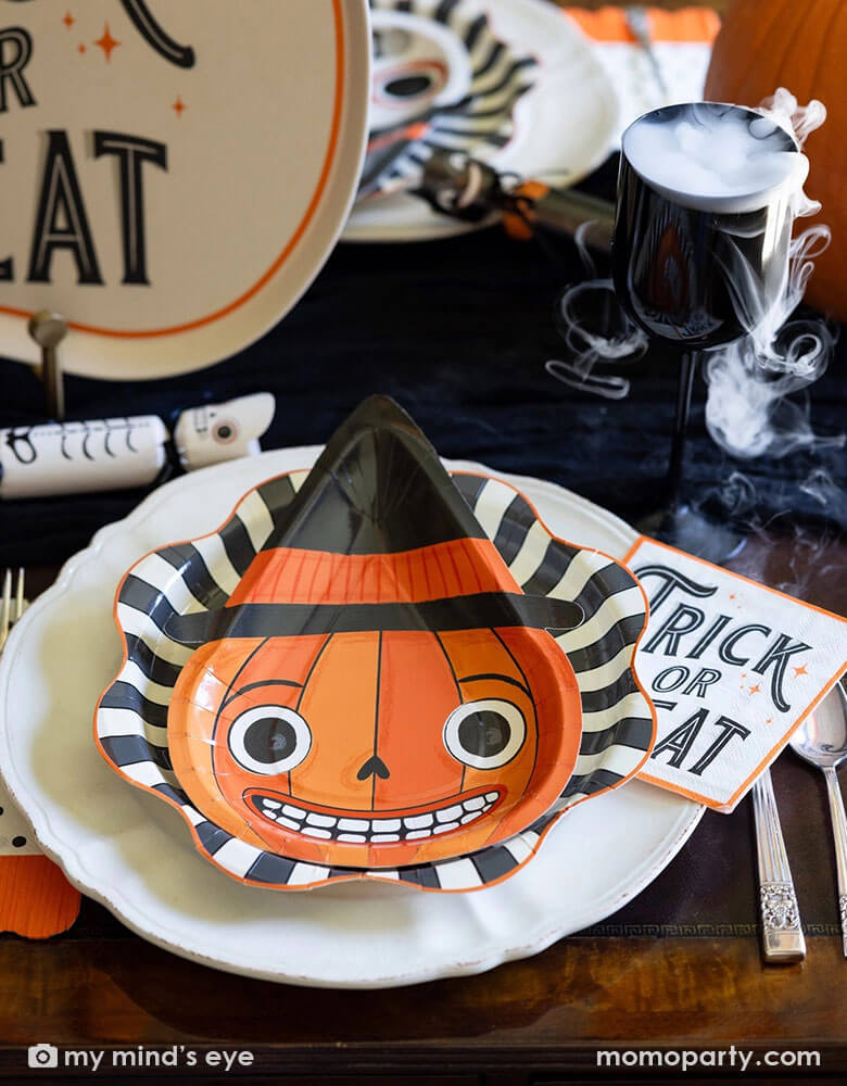 A vintage Halloween party table setting featuring vintage inspired Halloween party supplies from Momo Party including the vintage Halloween pumpkin shaped plate on the black starburst dinner plates and the vintage Halloween trick or treat napkins in vintage fonts. On the table there are some vintage Halloween party crackers in the design of a skeleton, a black cat and a pumpkin. There's also a black wine glass with dry ice as if there's magic smoke coming out for this spooky Halloween celebration!