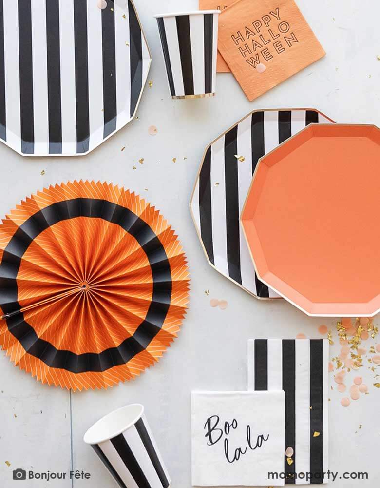 A table full of modern Halloween party supplies in the classic Halloween colors of orange and black from Momo Party including black and white striped plates, party cups and guest napkins, paired with orange plate, "Happy Halloween" small napkins and a paper party fan, with the black and white Boo La La small napkins and peach colored confetti around, this is a perfect inspo for a chic Halloween celebration.