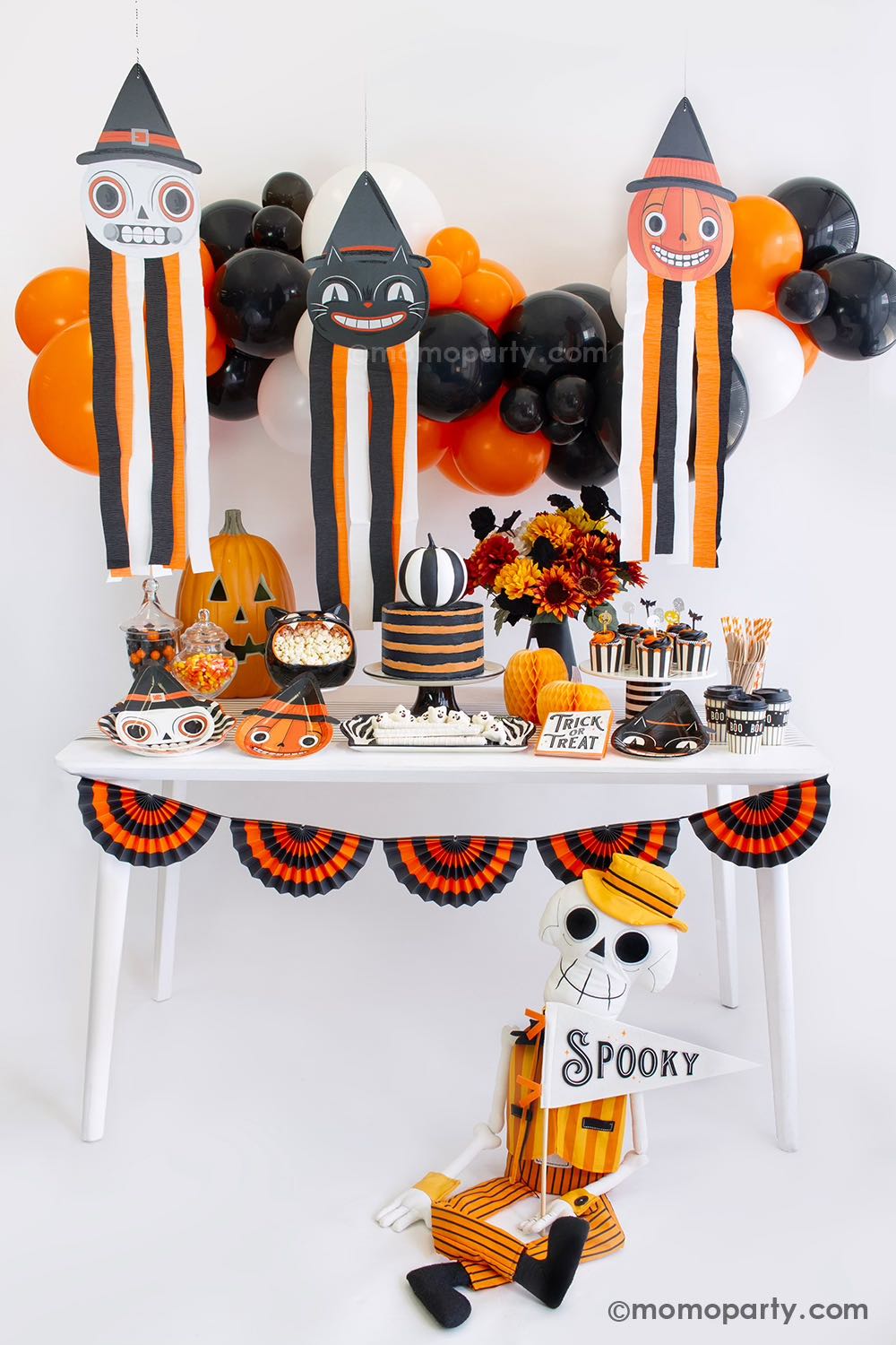 Momo Party's vintage Halloween party box displayed with classic themed decorations including black and orange hanging decorations with streamers, vintage-style tableware including pumpkin, skull and black cat shaped plates, striped party cups and trick or treat napkins, with a orange and black bunting garland and fun centerpieces like jack-o-lantern decoration and fall flower arrangement, it's perfect for a nostalgic Halloween celebration this spooky season.