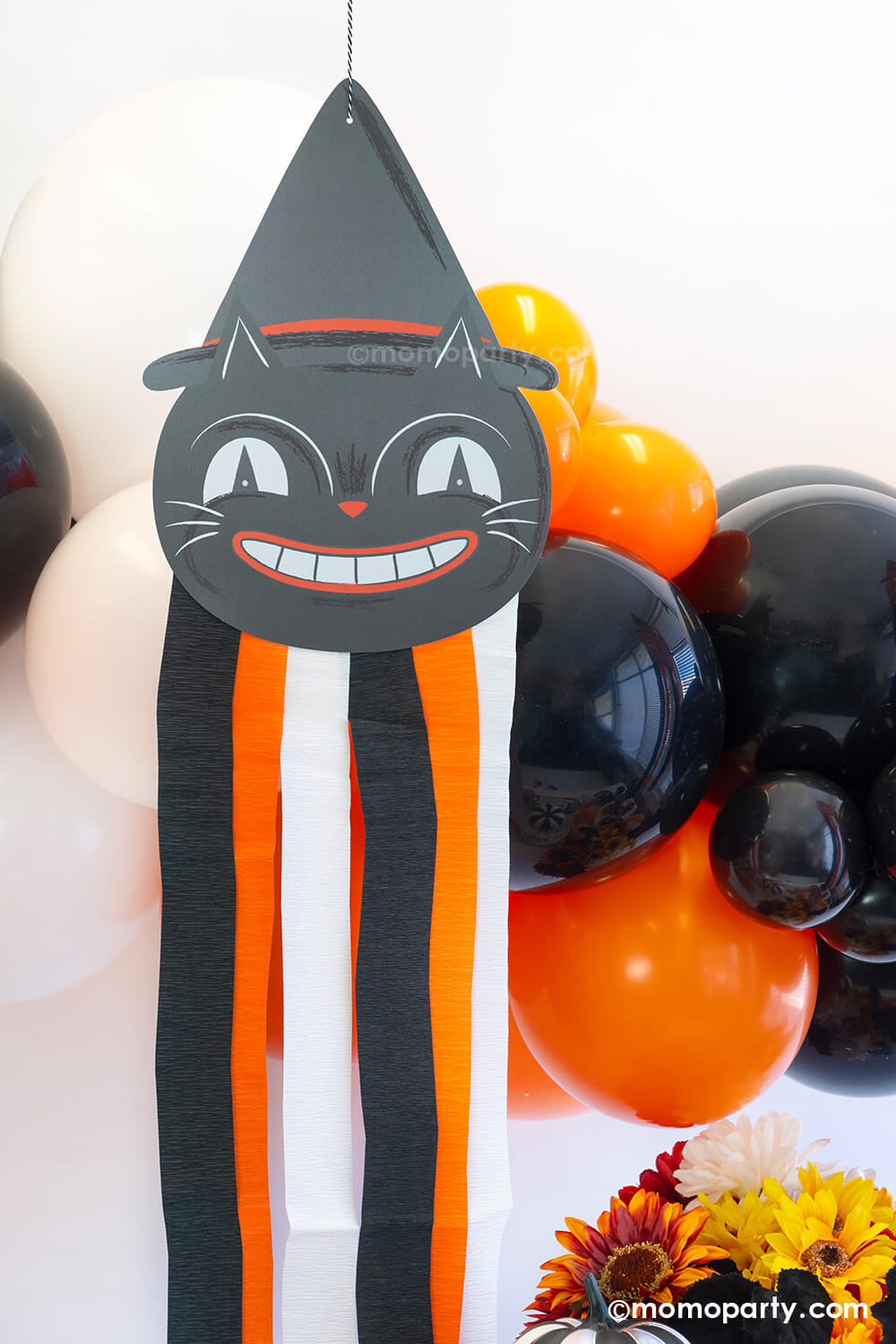 A close-up shot of a Vintage Halloween decorations featuring Momo Party's Halloween character black cat hanging decoration with streamers along with a balloon garland in classic Halloween colors of black, orange and white. On the day is a fall flower arrangement with matching colors, making this a great inspo for a simple and modern Halloween bash decorating idea that's also kid's friendly.