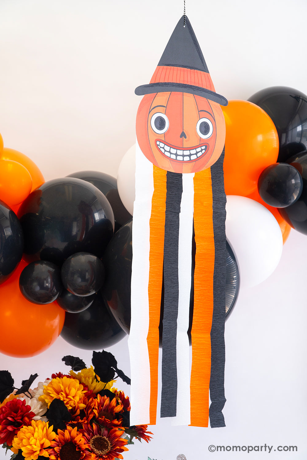 A close up shot of a Vintage Halloween decorations featuring Momo Party's Halloween character pumpkin hanging decoration with streamers along with a balloon garland in classic Halloween colors of black, orange and white. On the day is a fall flower arrangement with matching colors, making this a great inspo for a simple and modern Halloween bash decorating idea that's also kid's friendly.