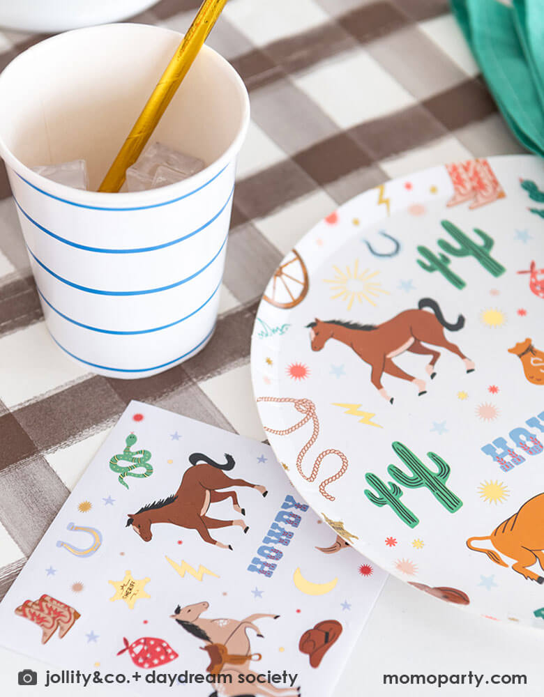 A closeup shot of a western cowboy themed party table tablescape featuring Momo Party's cowboy themed tableware by Jollity Co. and Daydream Society including the round cowboy icon patterned round plate, the small napkin and cactus honeycomb ., all on a brown gingham table runner, making this a modern yet fun tablescape for kid's rodeo, Western or cowboy themed birthday celebration. A perfect inpso for kid's "First Rodeo" first birthday celebration.  