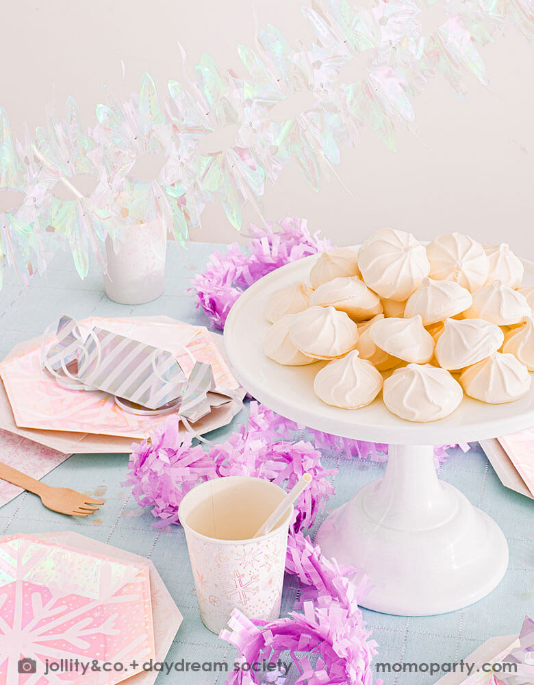 A whimsical winter wonderland or Frozen themed party table set up featuring Momo Party's frosted party collection including plates, napkins and party cups in snowflake design with iridescent accents. On the table there are some light pink meringue desserts and silver candy shaped treat boxes. With iridescent party garland above the table and lilac pink streamers on the table, this makes a great inspiration for a girl's dreamy winter themed celebration.