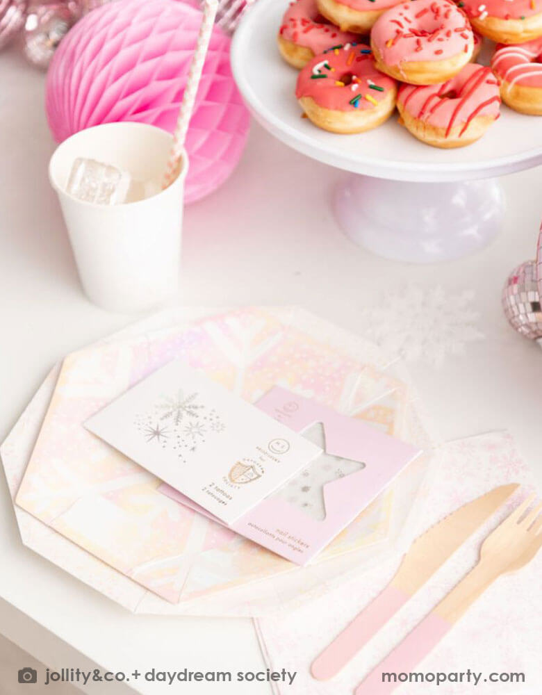 A close up of a whimsical winter wonderland or Frozen themed party table place set up featuring Momo Party's frosted party collection including plates, napkins, party cups, temporary tattoos and nail stickers in snowflake design with iridescent accents. On the table there are some mini donuts in pink color and some pink honeycombs and disco balls as decorations - makes this a great inspiration for a girl's dreamy winter themed celebration.