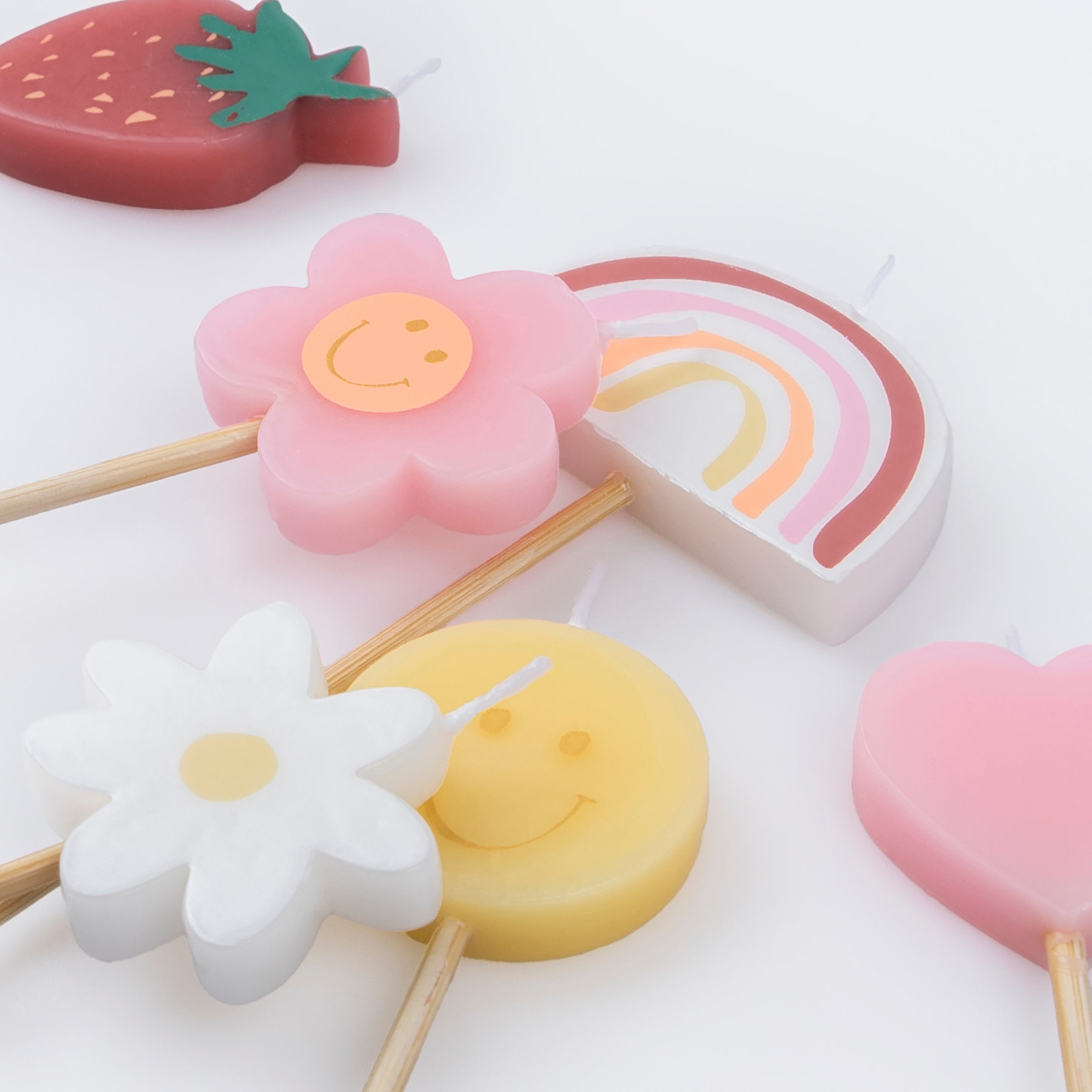 Details of Momo Party's Happy Face Icons Candles by Meri Meri. Featuring a set of 6 strawberry, a flower, a daisy flower, a rainbow, a smiley face, and a heart candle with long sticks, these colorful candles are perfect for any happy celebration!