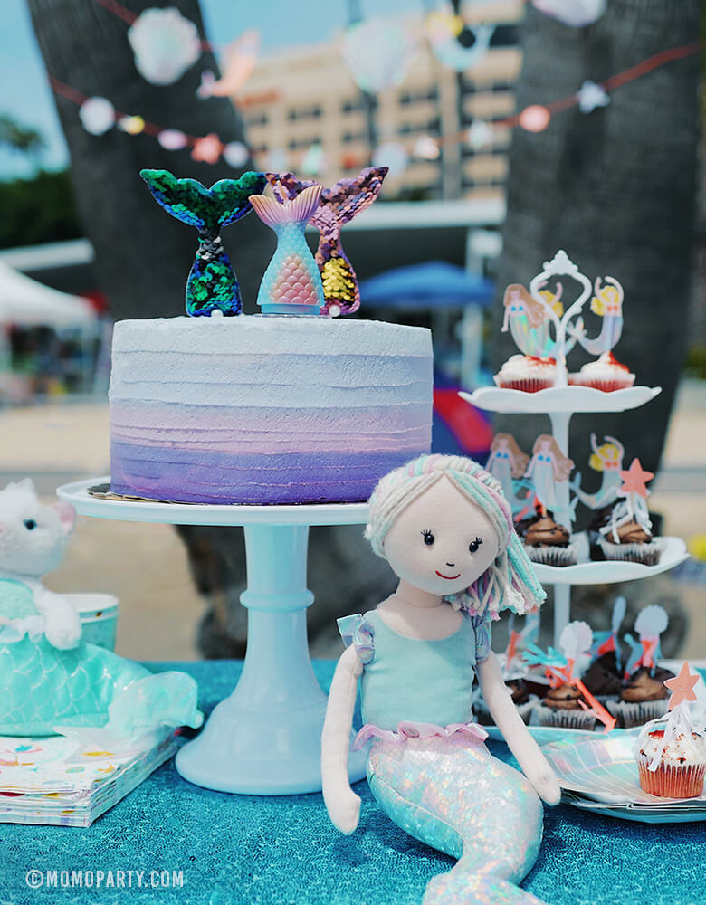 Mermaid beach party table set up with Pastel Ombre color Mermaid Tail Candle and reversible sequin fabric mermaid tail toppers on the cake, Jellycat Aqua-Lily Mermaid Doll, cupcakes